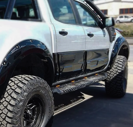 A lifted, black-and-white pickup truck is parked on a road, featuring large off-road tires and side steps with orange reflectors. It’s set against a blurred background.