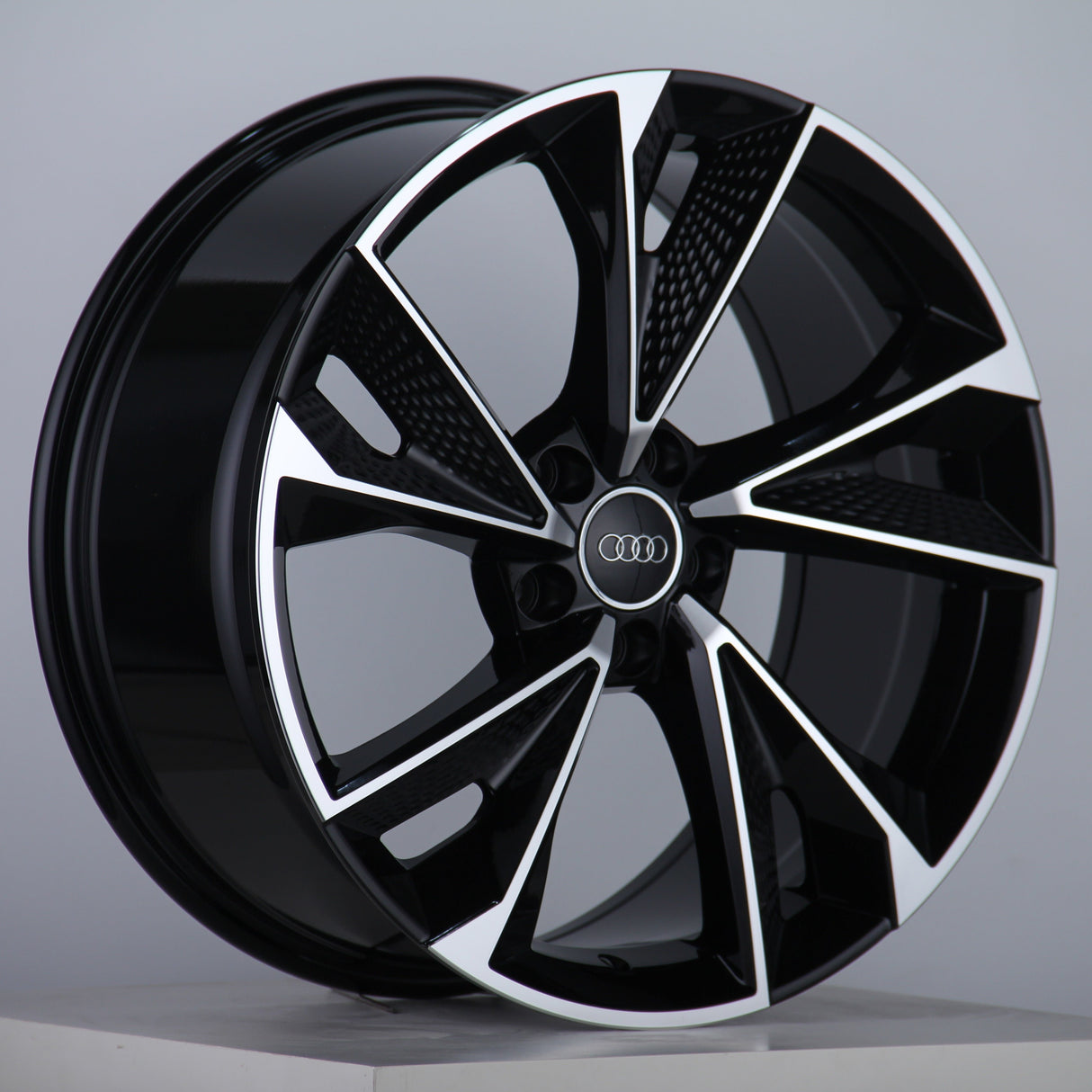 A black and silver alloy wheel displays its intricate, angular design with five spokes. The Audi logo is centered. It is set against a plain gray background, resting on a white surface.