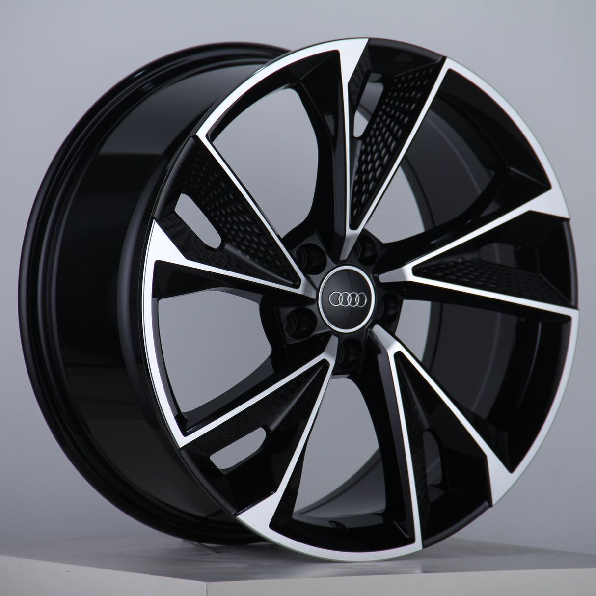A glossy, black alloy wheel features silver accents and the Audi logo at the center, displayed on a neutral gray background with a studio setting.