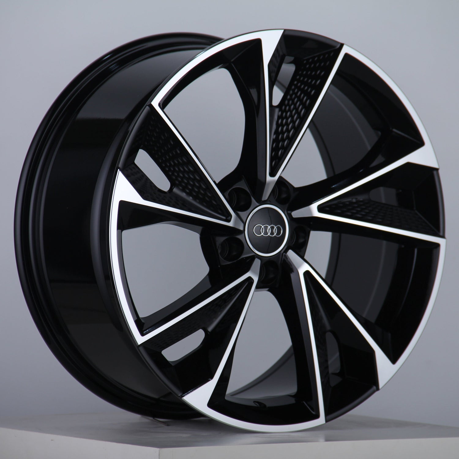 A glossy, black alloy wheel features silver accents and the Audi logo at the center, displayed on a neutral gray background with a studio setting.
