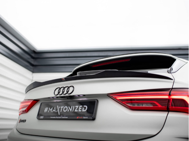 Car rear displays "#MAXTONIZED" on a license plate. The white Audi, featuring a spoiler and red taillights, is parked indoors beside gray and white paneling.