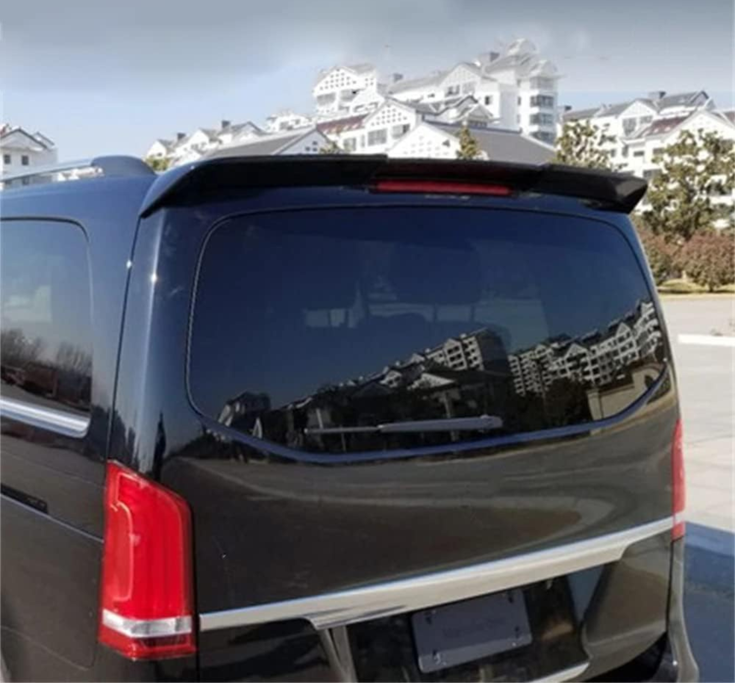 A black vehicle with tinted windows is parked, reflecting nearby white buildings with sloped roofs. The scene is set in a sunny, open outdoor area.