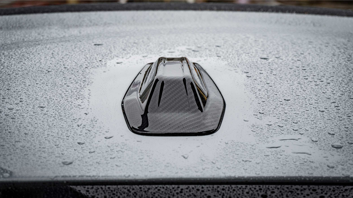 A black, shark fin car antenna rests on a gray, wet car roof, with raindrops scattered across the surface creating a textured appearance.