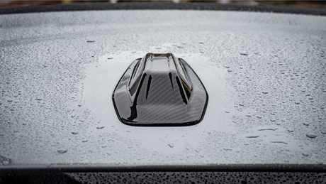 A black, shark fin car antenna rests on a gray, wet car roof, with raindrops scattered across the surface creating a textured appearance.