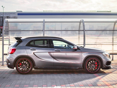 A silver sports car with red-accented wheels is parked on a tiled lot in front of a clear shelter. The background includes building signage and a cloudy sky.