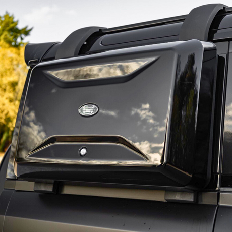 A sleek black storage box is mounted on a vehicle's roof, reflecting clouds in a clear sky. An emblem with text "FORCE" is displayed on its front. Trees in the background.