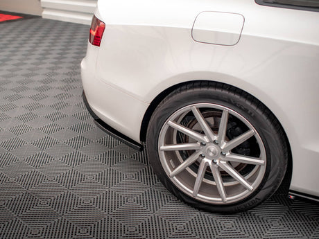 Car rear wheel is parked on a textured gray garage floor. The white vehicle's body features a visible fuel cap, with red taillight on display in the upper corner.