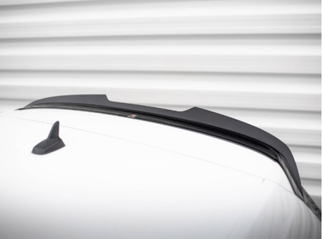 A sleek black car spoiler is mounted on a white vehicle roof, with a small shark-fin antenna nearby, set against a blurred horizontal striped background.