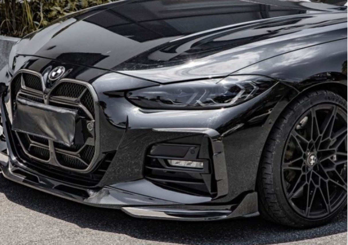 A sleek, black car displays its front grille and headlight assembly while parked on an outdoor surface. The wheel features a detailed spoke design, with greenery visible in the background.