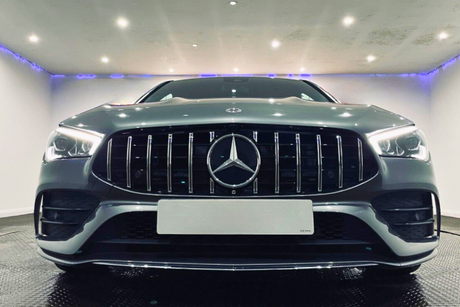 A silver car with a prominent grille and illuminated headlights is parked indoors under bright ceiling lights, creating a sleek and modern atmosphere. A Mercedes-Benz logo is centered on the grille.