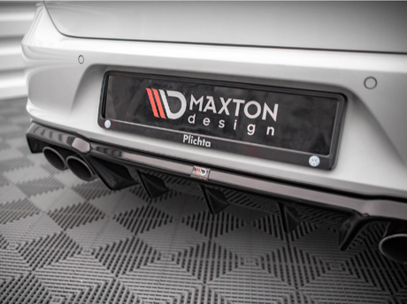 A car bumper features a license plate that reads "MAXTON design Plichta." Dual exhaust pipes are visible beneath. The car rests on a textured, patterned floor in a well-lit showroom.