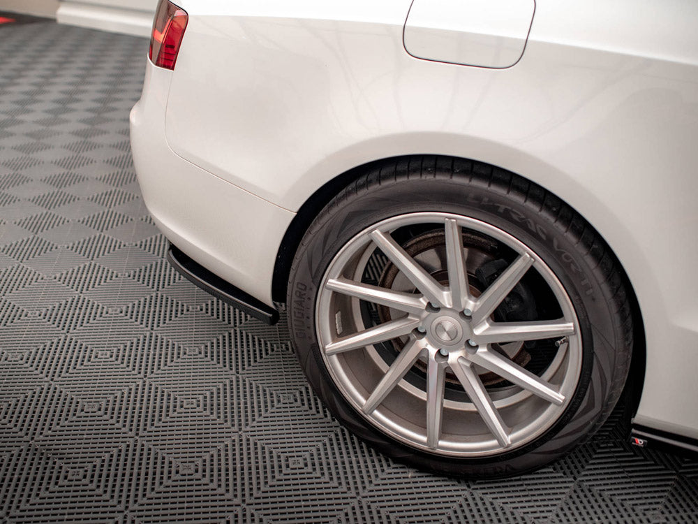 A white car's rear wheel with a silver alloy rim is stationary on a textured gray garage floor, showing partial tail light and fuel door.