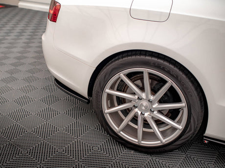 A white car's rear wheel with a silver alloy rim is stationary on a textured gray garage floor, showing partial tail light and fuel door.