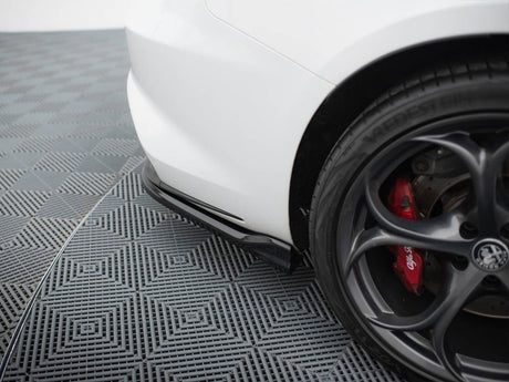 A white car's rear bumper is parked on a patterned grid floor. The black alloy wheel features red brake calipers labeled "Alfa Romeo." The tire brand reads "VREDESTEIN."