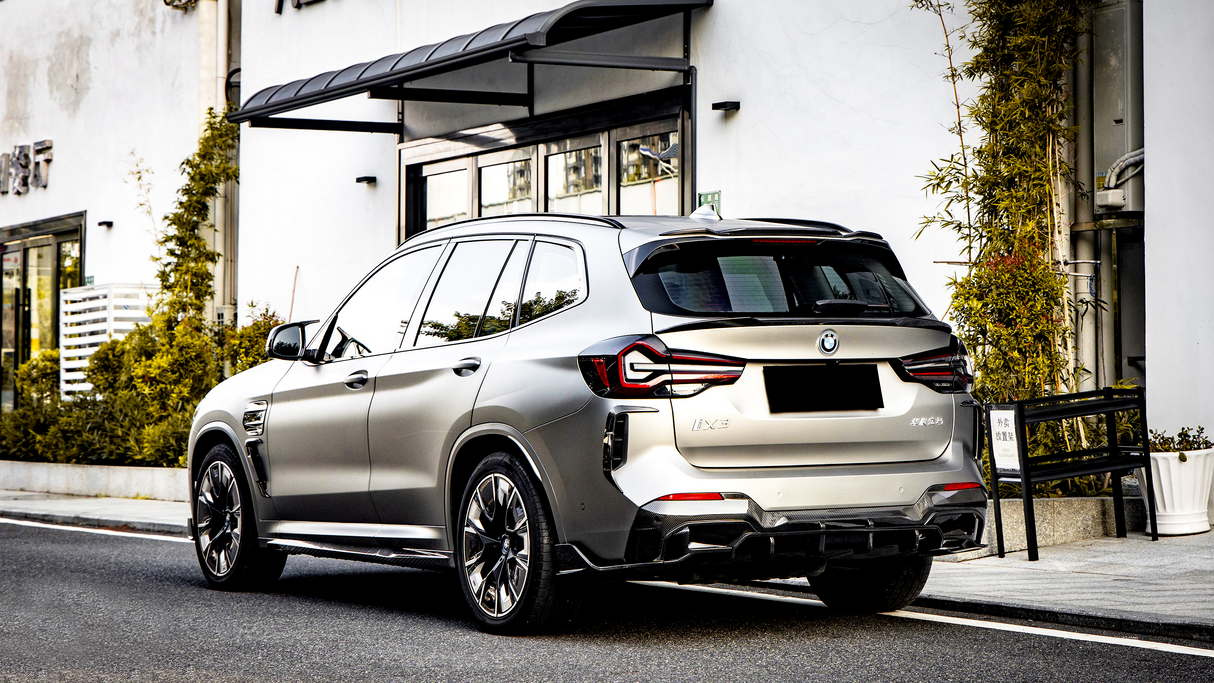 A metallic BMW iX3 parked on a street beside a building with glass doors and greenery. The rear of the car shows sleek taillights and "iX3" and other text on the back.