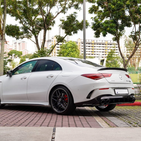White Mercedes-Benz CLA 45 AMG parked on a patterned pavement, surrounded by trees and buildings in the background. The rear of the car features distinctive taillights and quad exhaust pipes.
