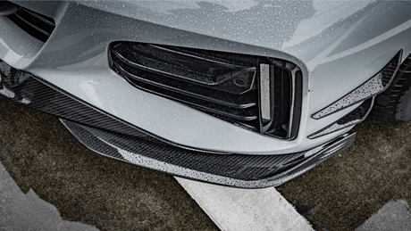 A car's front bumper, featuring sleek black grills and carbon fiber accents, is covered in rain droplets, situated on a wet asphalt surface with white road markings.