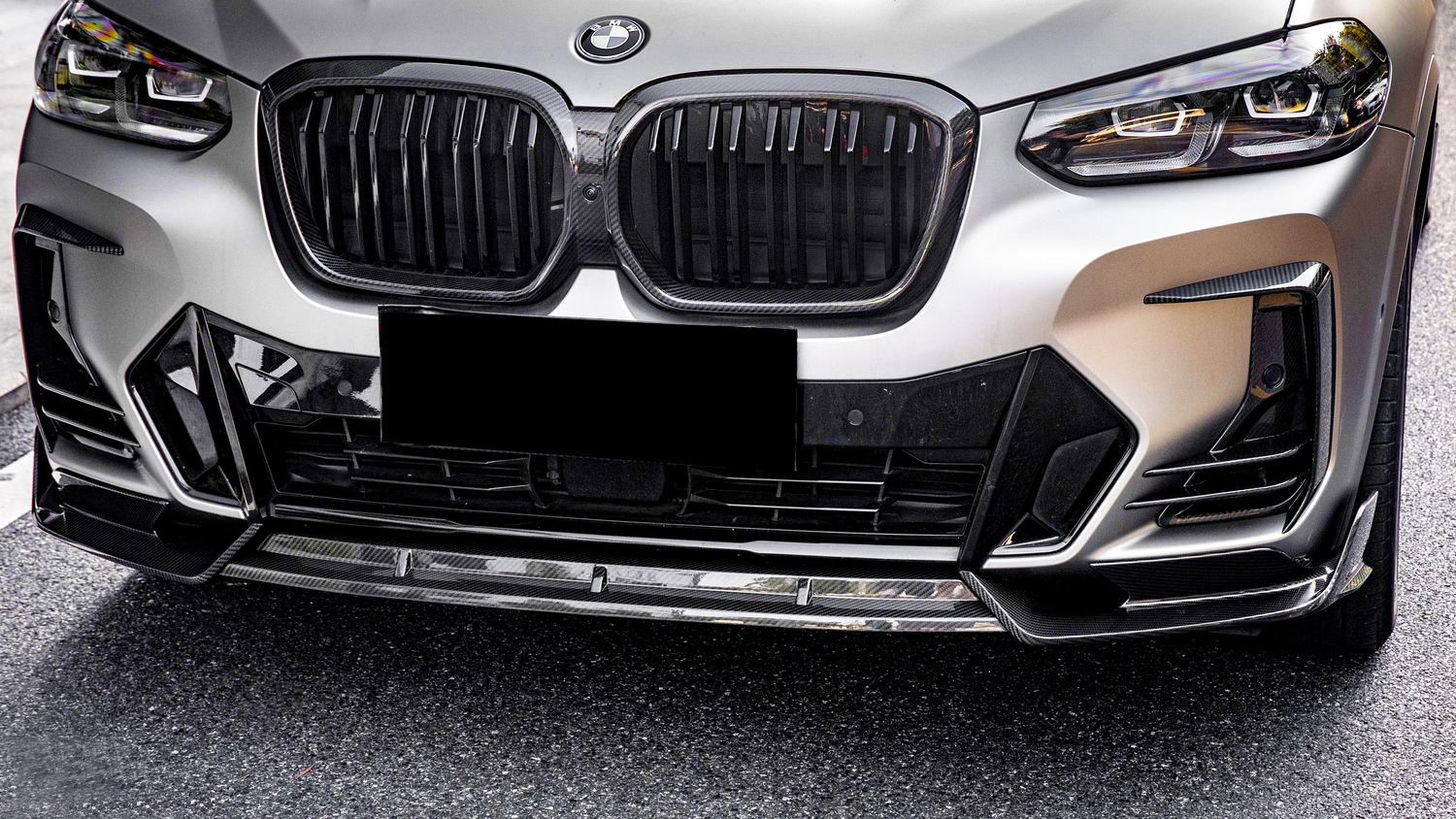 A BMW car front features a prominent grille and sleek headlights, parked on a textured asphalt surface. A black license plate obscures the lower section.