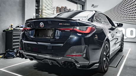 A black BMW M425i car is parked indoors, reflecting light on its glossy surface. Nearby, a tool cabinet and bottle indicate a workshop environment. "DryCarbon" branding is visible on the wall.