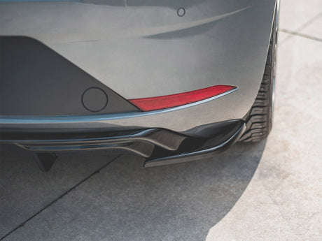 A car's rear bumper with a red reflector is parked on a sunlit, textured concrete surface, highlighting the curvature and tire tread.