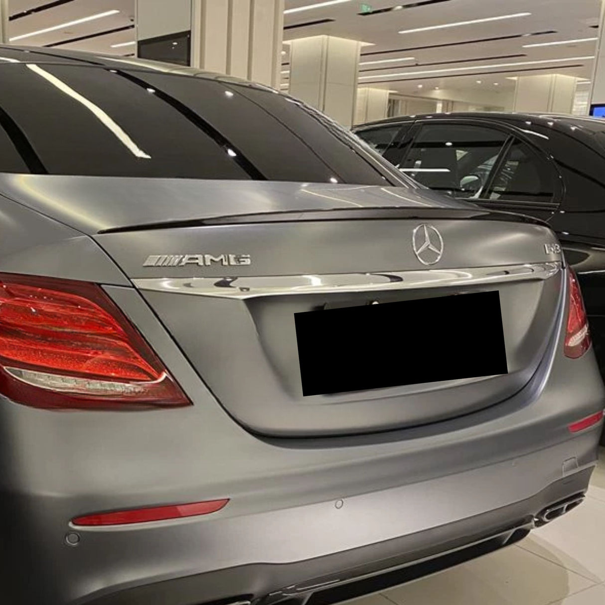 A silver Mercedes AMG E43 is parked indoors, showcasing badges on the trunk. The surrounding area has a modern, well-lit showroom with another vehicle visible nearby.