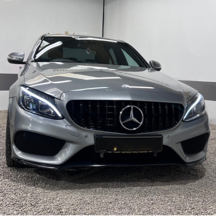 A silver car with illuminated headlights is parked indoors on a gravel floor, featuring a prominent emblem on the grille and reflective lighting in a simple white-walled setting.
