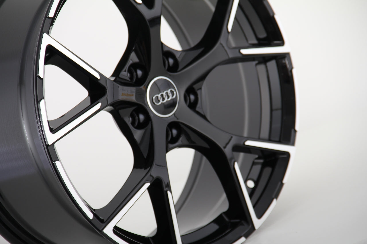 A glossy black alloy wheel with a silver trim has the Audi logo in the center, displayed against a plain, neutral background.