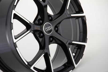 A glossy black alloy wheel with a silver trim has the Audi logo in the center, displayed against a plain, neutral background.