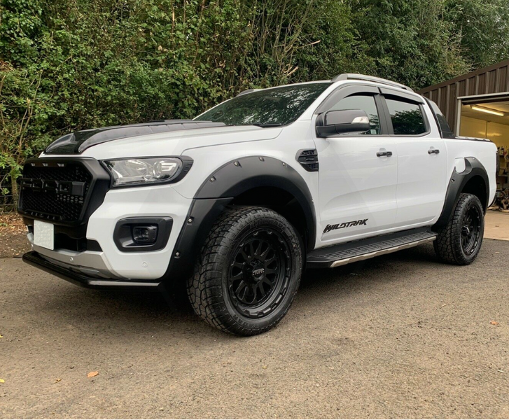 A white pickup truck with black trim and large tires is parked on a paved area near a building. The truck has "WILDTRAK" written on its side, surrounded by green foliage.