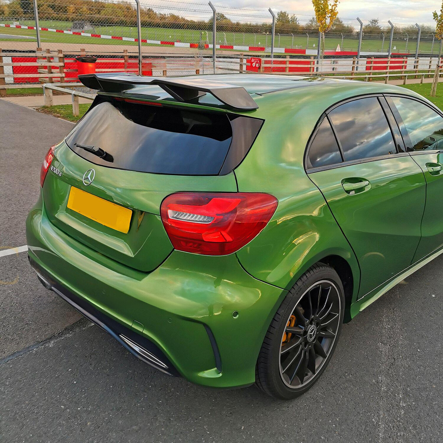 A green Mercedes-Benz hatchback is parked on a racetrack's asphalt surface. It's positioned near fencing and a grassy area, featuring a rear spoiler and tinted windows.