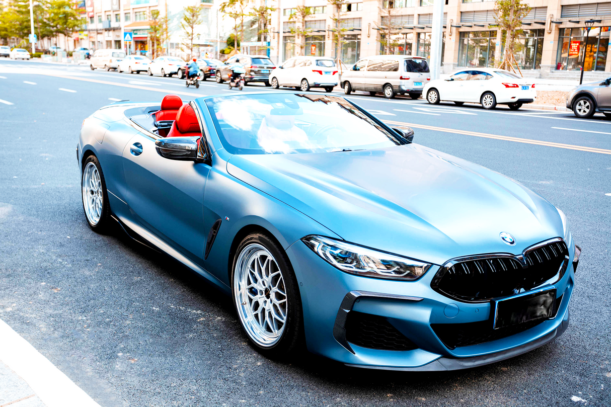 A blue convertible car with red seats is parked on a city street, surrounded by traffic and buildings, under a clear sky.