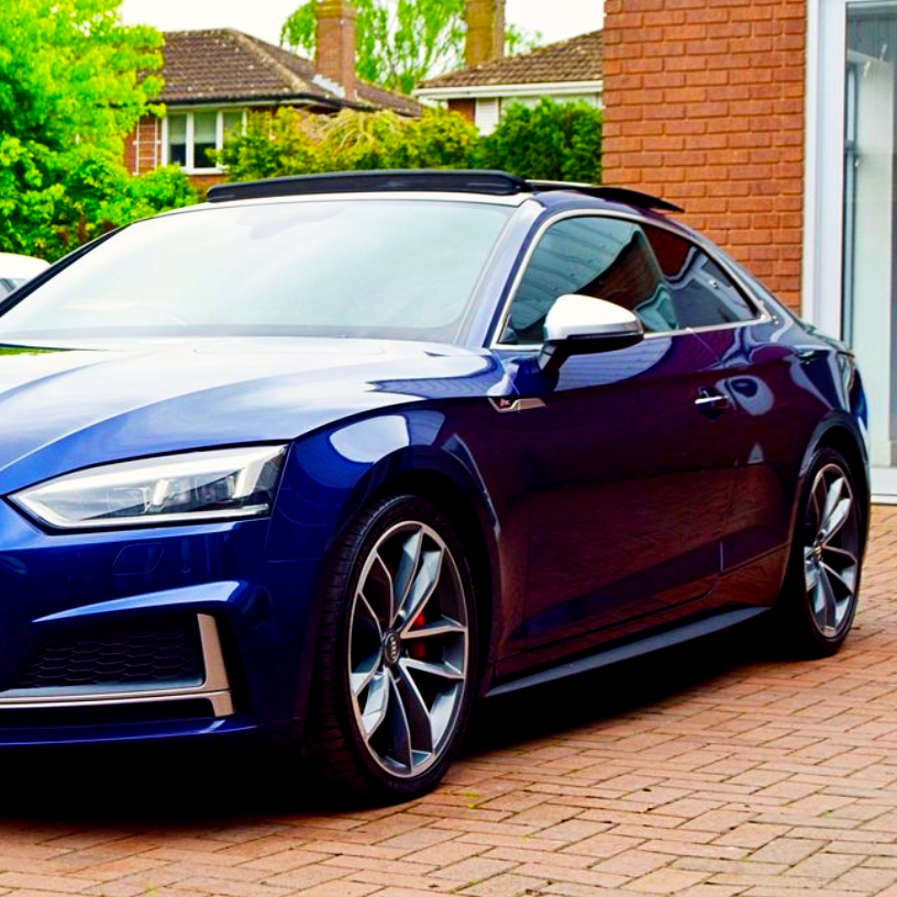 A blue sports car is parked on a brick driveway with houses and greenery in the background. The car has sleek lines and a sunroof is partially visible.