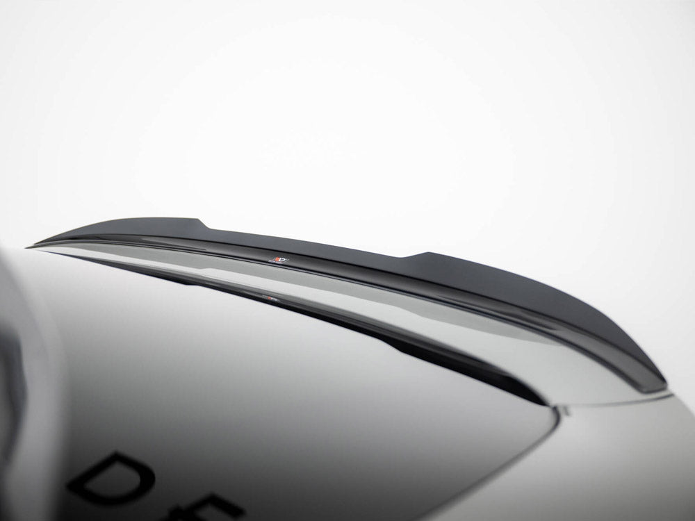 A black car roof spoiler is mounted on a smooth, grey vehicle surface. Partial letters "DE" are visible on the car, set against a plain, white background.