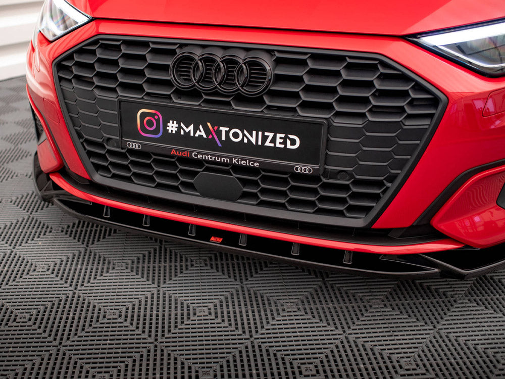A red car's front grille dominates, with a black license plate reading "#MAXTONIZED Audi Centrum Kielce" against a textured black floor in a showroom setting.