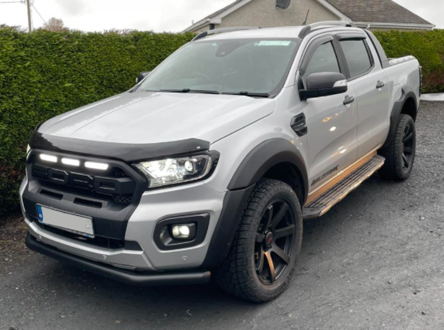 A silver pickup truck, stationary, is parked on a gravel driveway. It features a custom grille with lights and is surrounded by green hedges and a house in the background.