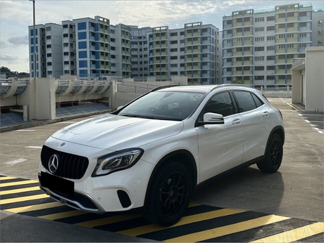 A white SUV with tinted windows is parked on a rooftop near black and yellow stripes. In the background, tall residential buildings stand against a partly cloudy sky.