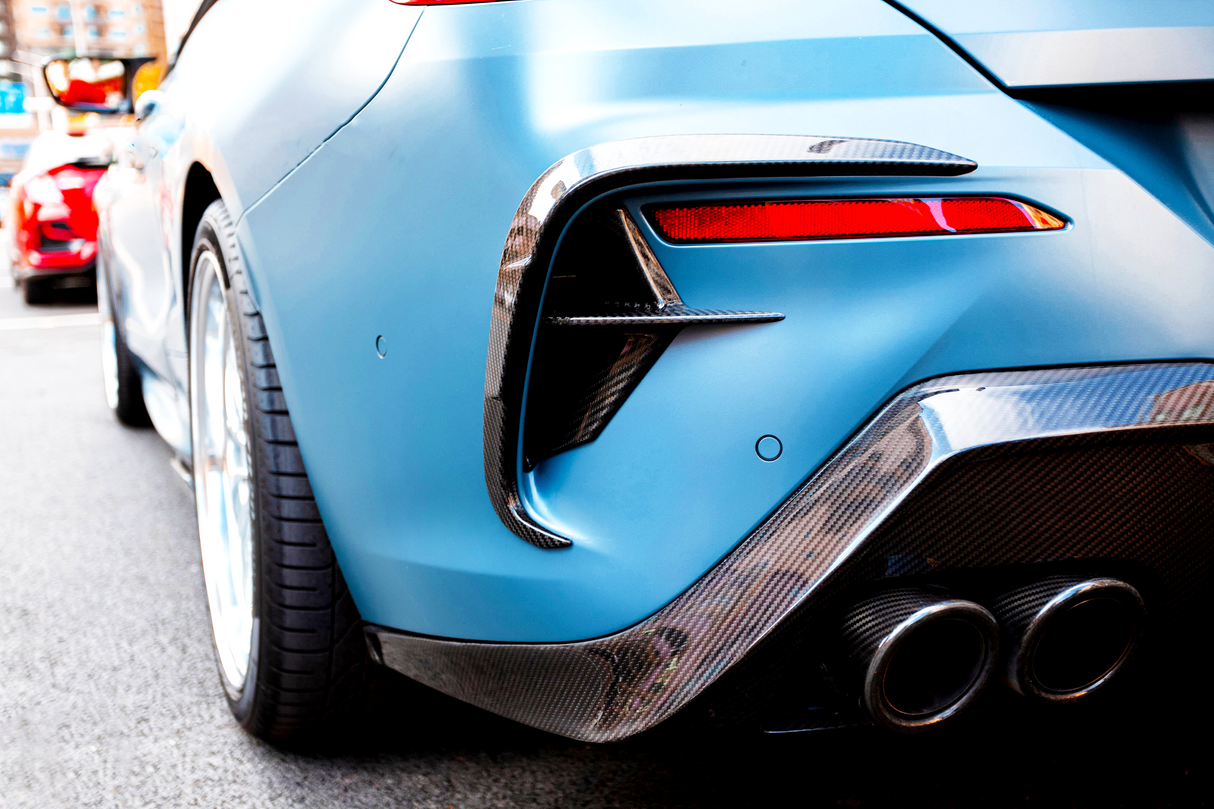 A blue sports car's rear features chrome exhausts and red tail lights, parked on a city street with another car partially visible in the background.
