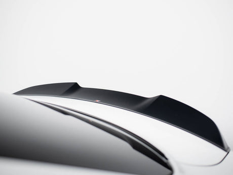 A black car spoiler is mounted on a sleek, white vehicle in a minimalistic setting. The background is a plain, light gray color, emphasizing the spoiler's contours.