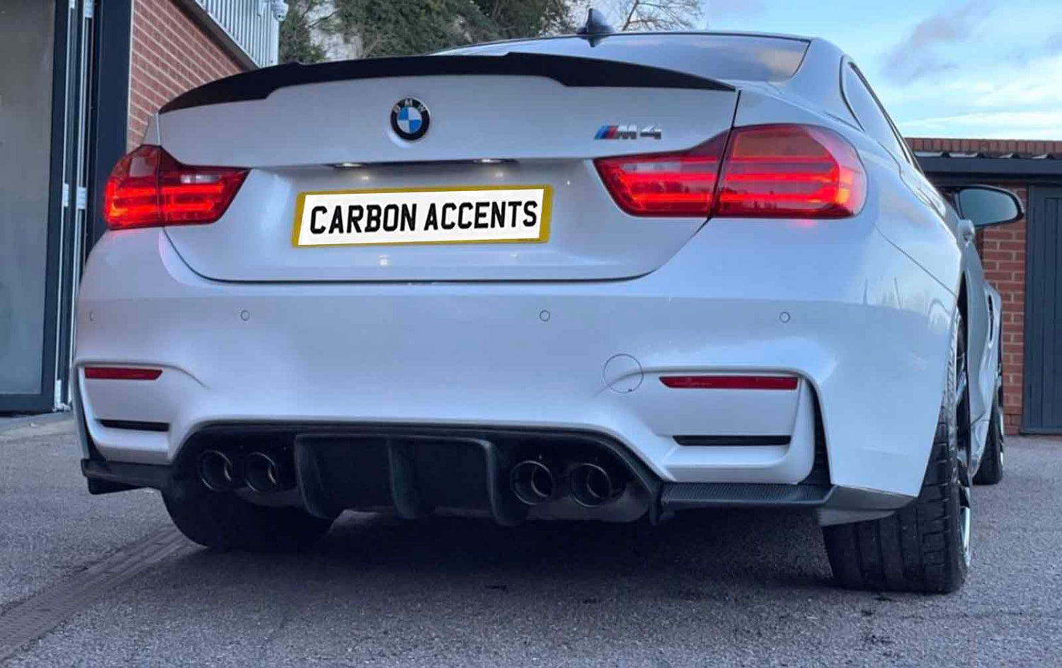 A white BMW M4 is parked, featuring dual exhausts and illuminated rear lights. The license plate reads "CARBON ACCENTS." Brick walls and a building are visible in the background.