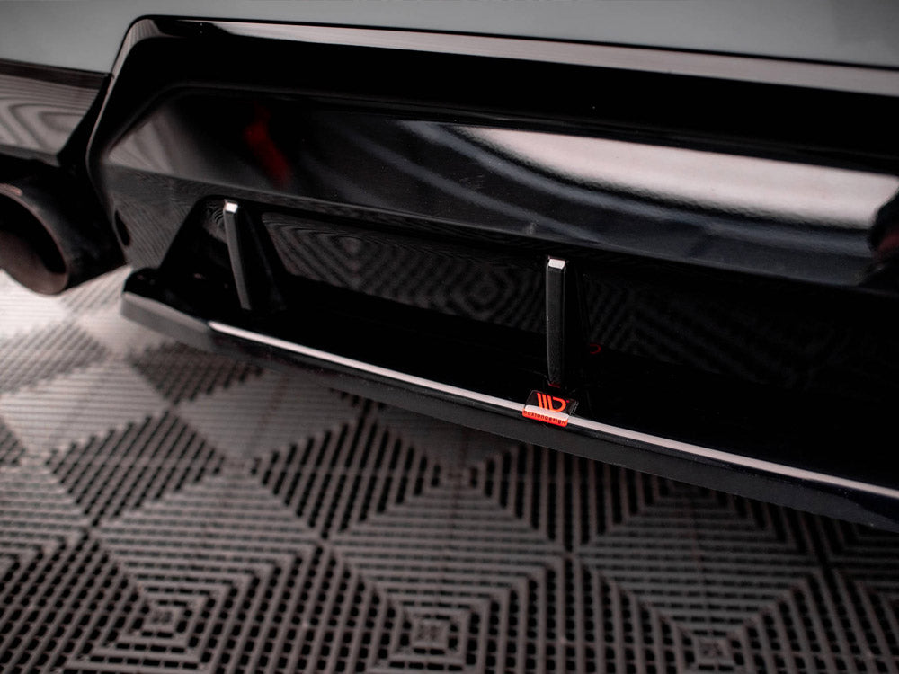 A black car's rear bumper features an aerodynamic diffuser with two vertical fins and a red logo. It rests on a patterned rubber matting surface in an indoor space.