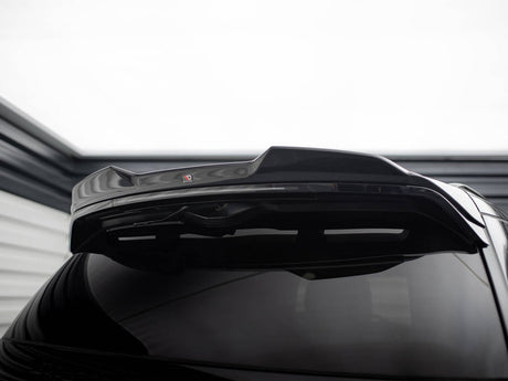 A shiny black car's rear spoiler is raised, highlighting its glossy finish. In a dimly lit garage setting, a small red and black "V" logo is visible on the spoiler.
