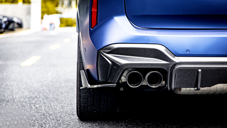 A blue car's rear with dual exhaust pipes and carbon fiber detailing is stationary on a paved road; blurred background suggests an urban environment.