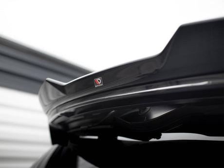 A black car spoiler features a "Vortex Generator" sticker, mounted on the vehicle's trunk. The background includes blurred, horizontal lines of a light-colored structure, suggesting an industrial environment.