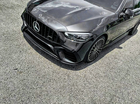 A sleek, dark-colored Mercedes car is parked on a gravel surface, highlighting its prominent grille and intricate wheel design under a partly cloudy sky.