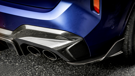 A car's blue rear bumper features a glossy carbon fiber diffuser with three exhaust pipes, parked on an asphalt surface. The setting suggests a modern automotive design.