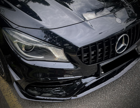 A black car's front end with a prominent grille and headlight is parked on a street, reflecting trees and clouds, featuring a three-pointed star emblem on the hood.