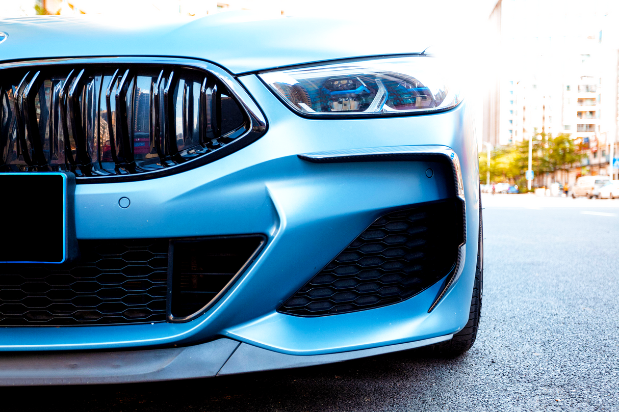 A sleek car grille and headlight on a blue vehicle, positioned on a city street with buildings and trees in the background.