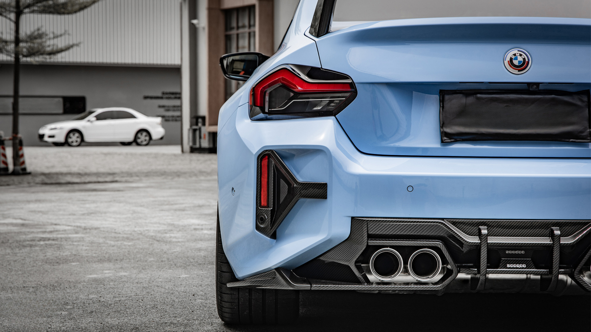 A blue car's rear is parked in a lot, featuring prominent exhausts and taillights, with a white car and industrial building in the blurred background.