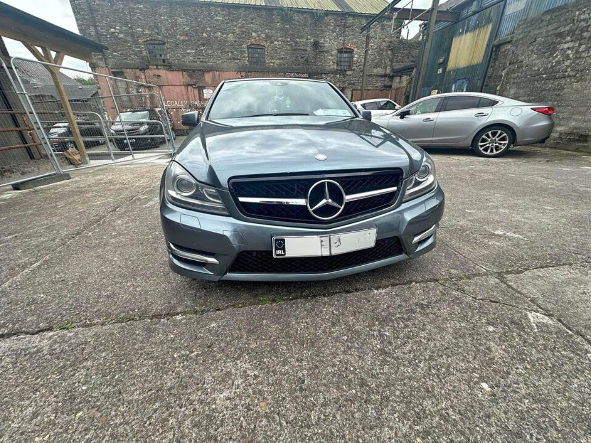 A silver Mercedes-Benz parked on a concrete surface, surrounded by several other vehicles, near a weathered building and wire fencing. License plates show symbols but no legible text.