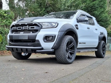 A white Ford pickup truck with "WILDTRAK" branding stands stationary on a paved surface. It features large tires and black detailing, surrounded by a leafy, wooded area.